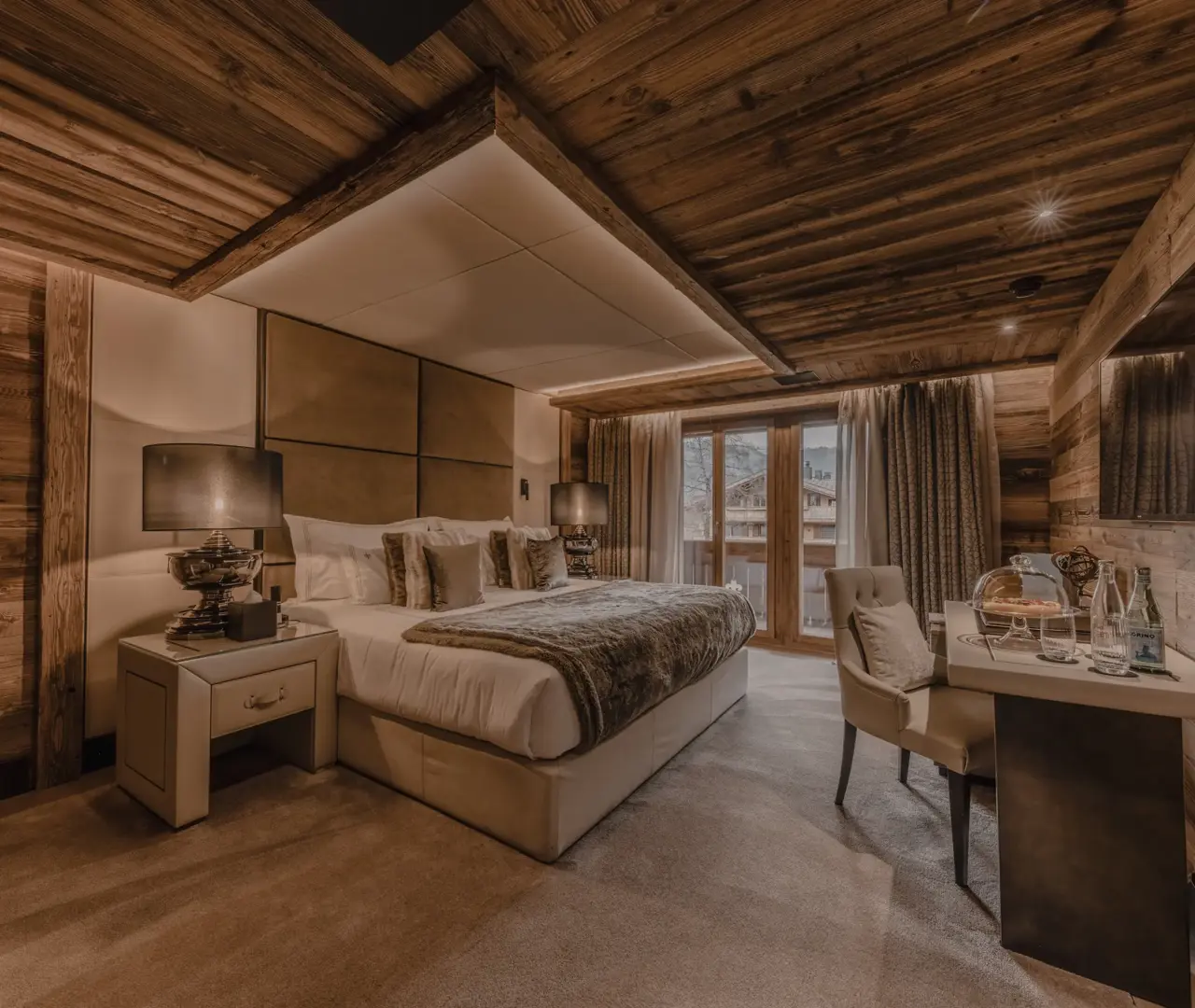 A cozy luxury chalet bedroom with warm lighting, wooden walls and ceiling beams, a large bed with plush pillows and fur accents, bedside lamps, and a small desk setup by the window overlooking the mountains.