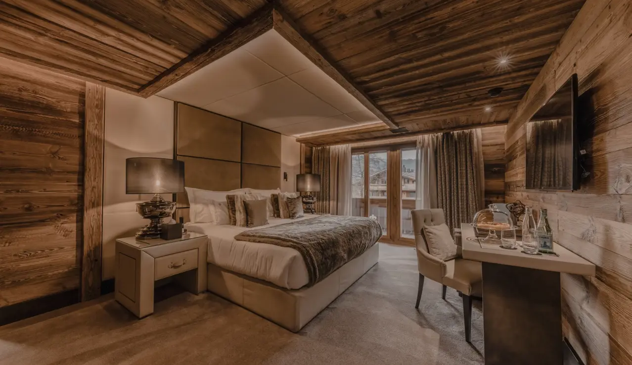 A cozy luxury chalet bedroom with warm lighting, wooden walls and ceiling beams, a large bed with plush pillows and fur accents, bedside lamps, and a small desk setup by the window overlooking the mountains.
