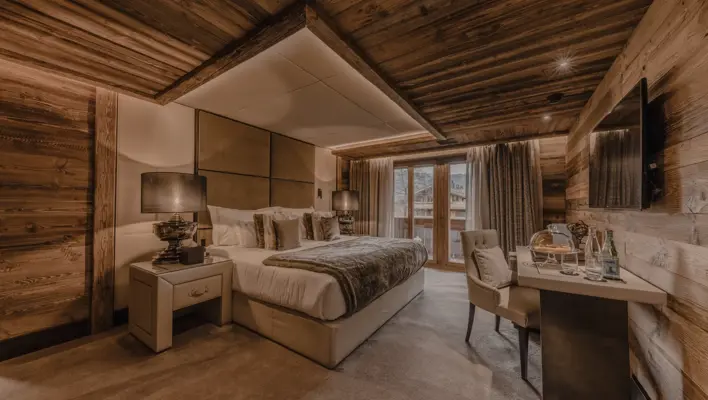 A cozy luxury chalet bedroom with warm lighting, wooden walls and ceiling beams, a large bed with plush pillows and fur accents, bedside lamps, and a small desk setup by the window overlooking the mountains.