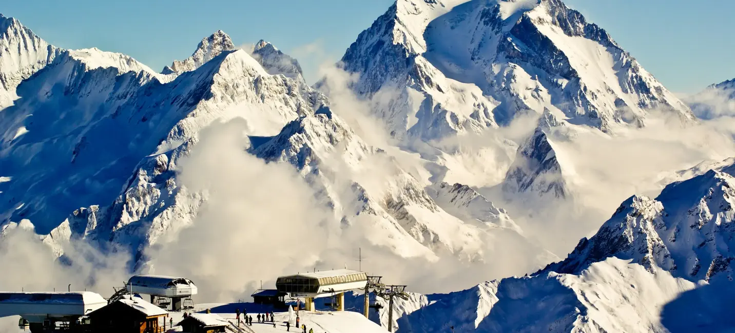 جبال الألب المغطاة بالثلوج تحت سماء زرقاء صافية مع محطة مصعد تزلج ومتزلجين يظهرون على المنحدر في المقدمة.