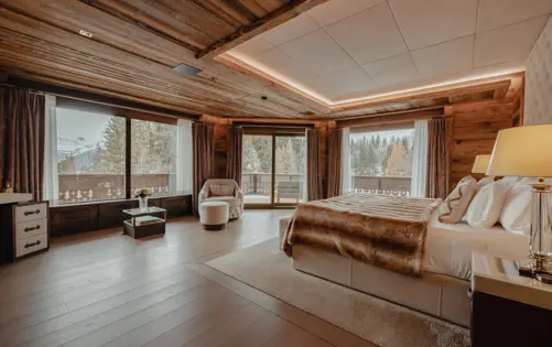 Grande chambre principale d’un chalet alpin de luxe avec vue panoramique sur les montagnes et la forêt, boiseries naturelles, éclairage doux et lit king-size habillé de fourrure et de lin pour une retraite montagnarde élégante.