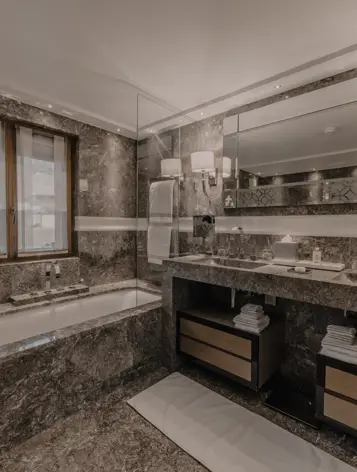 A luxurious bathroom featuring elegant marble walls and floors, a built-in bathtub by the window, and a wide vanity with dual sinks beneath a large mirror. Soft lighting and sleek fixtures create a sophisticated spa-like ambiance.