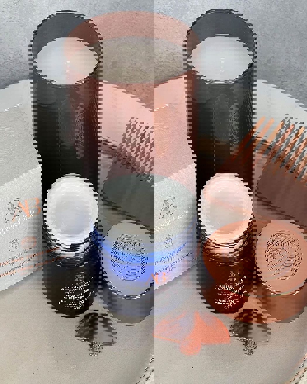 Pot ouvert du masque crème pour le visage Augustinus Bader avec couvercle cuivré, bougie et boîte présentés sur une surface en marbre.