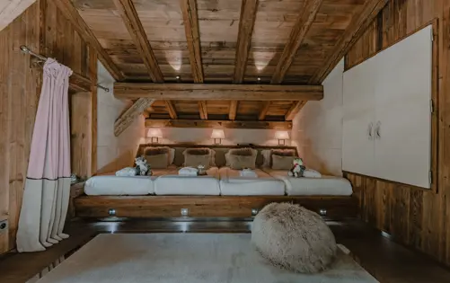 Уютная детская спальня в шале с деревянными балками, мягким освещением, плюшевыми игрушками и несколькими односпальными кроватями в тёплом альпийском стиле.