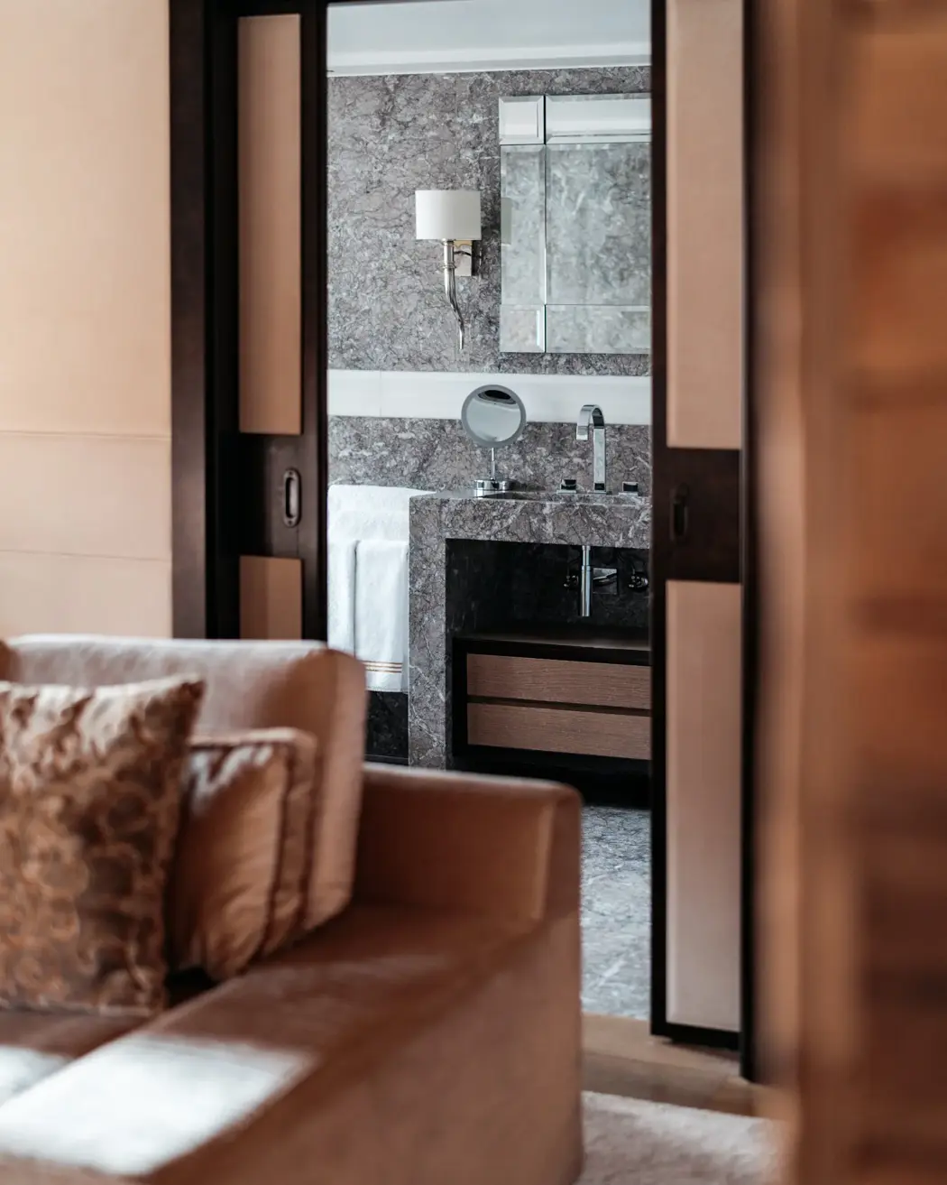 Luxurious view from a cozy living area into a marble bathroom, featuring a modern vanity with a mirror, elegant lighting, and sliding doors framing the scene.