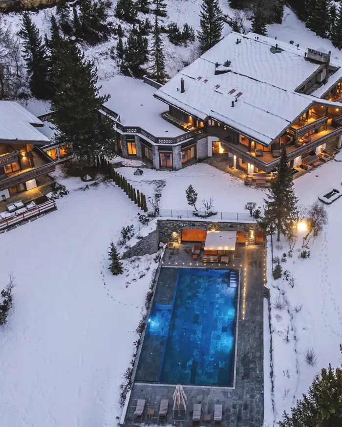 A luxury mountain chalet illuminated at dusk, surrounded by fresh snow and featuring a heated outdoor pool for an unforgettable alpine retreat.