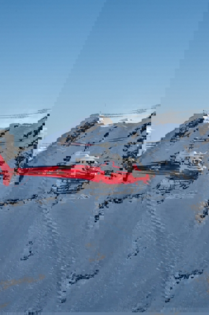 Red rescue helicopter flying over snowy Swiss Alps near a mountain station under a clear blue sky.