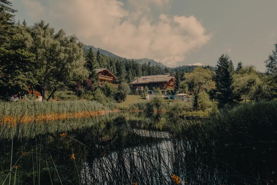 Chalets alpins au bord d’un lac, entourés de verdure luxuriante et de collines boisées, se reflétant dans l’eau calme sous un ciel d’été partiellement nuageux.