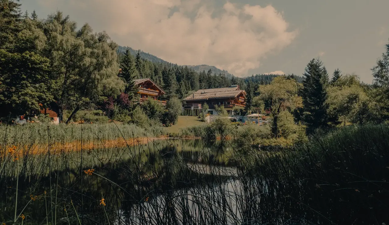 Lakeside alpine chalets surrounded by lush greenery and forested hills, reflecting in the calm water under a partly cloudy summer sky.