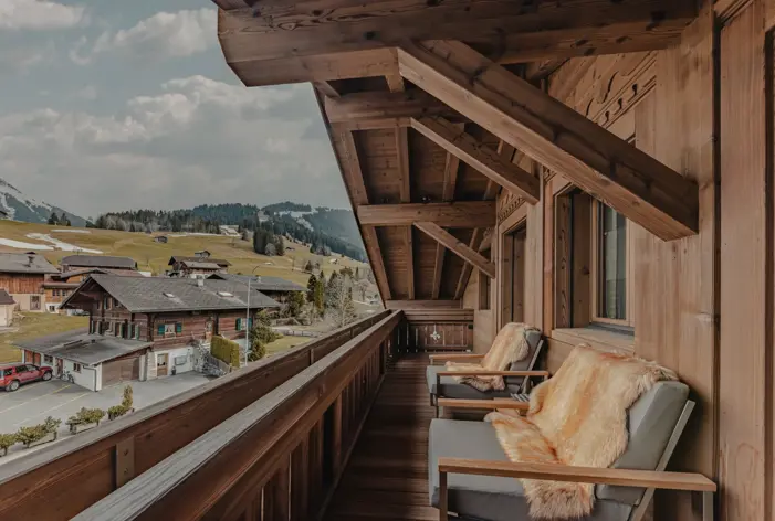 Warm wooden balcony with fur-covered chairs overlooking a picturesque alpine village, surrounded by rolling green hills and patches of melting snow under a cloudy sky.