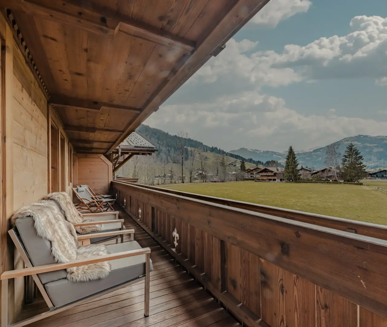 A cozy wooden balcony with fur-draped chairs overlooking a green alpine meadow and distant chalets, set against a backdrop of forested hills and a partly cloudy sky.
