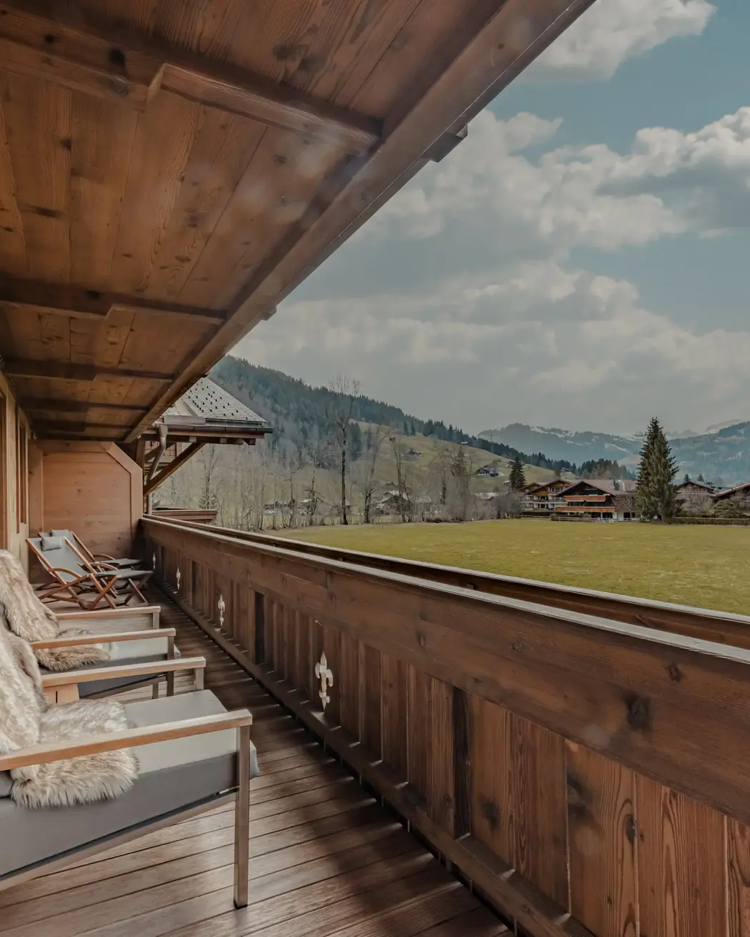 A cozy wooden balcony with fur-draped chairs overlooking a green alpine meadow and distant chalets, set against a backdrop of forested hills and a partly cloudy sky.