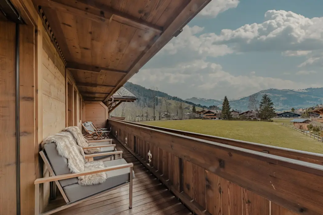 Balcon en bois chaleureux avec des chaises recouvertes de fourrure, surplombant une prairie alpine verte et des chalets au loin, avec en arrière-plan des collines boisées et un ciel partiellement nuageux.