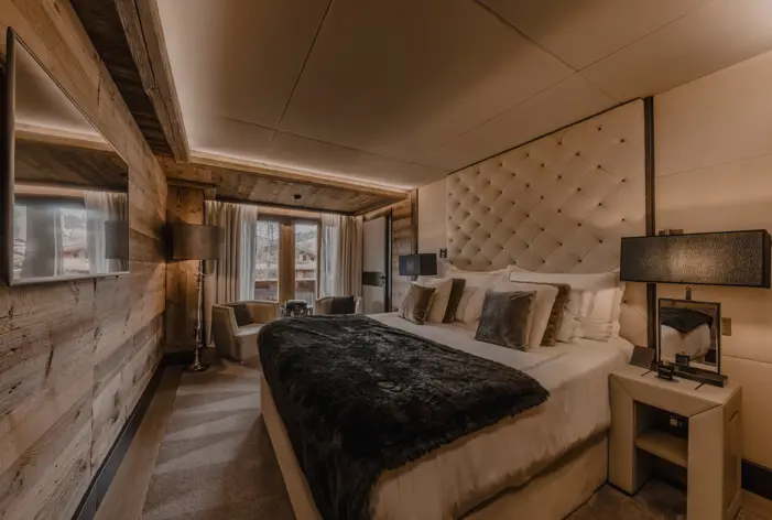 Elegant chalet-style bedroom featuring a large, plush bed with a tufted headboard, soft lighting, and cozy decor in warm neutral tones, with wooden walls and a view of the snowy mountains through glass doors.