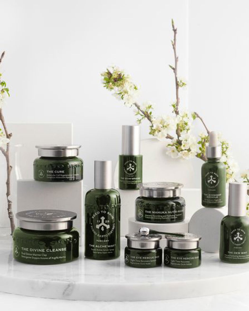 Présentation élégante de la collection de soins Seed to Skin avec des flacons et pots verts disposés sur une surface en marbre ornée de fleurs blanches.