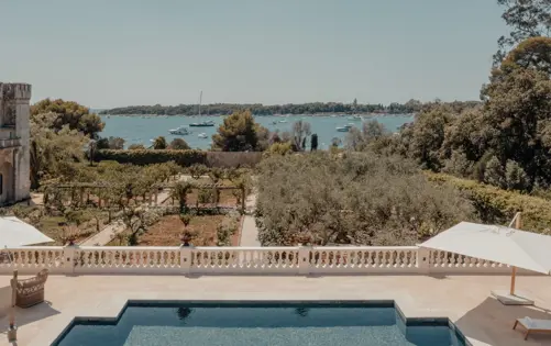 Piscine d’une villa de luxe surplombant un jardin méditerranéen et la mer bleue, avec des yachts ancrés au loin sous un ciel d’été clair.