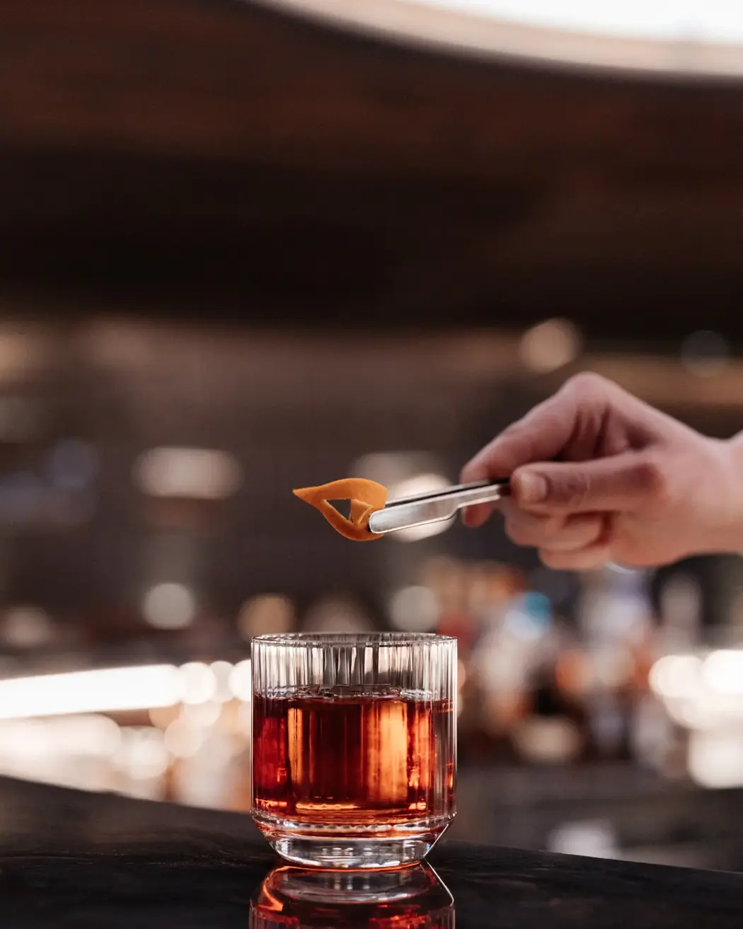 A close-up of a hand using tongs to place an orange peel garnish onto a glass of amber-colored cocktail, set on a dark bar counter with a softly lit background.