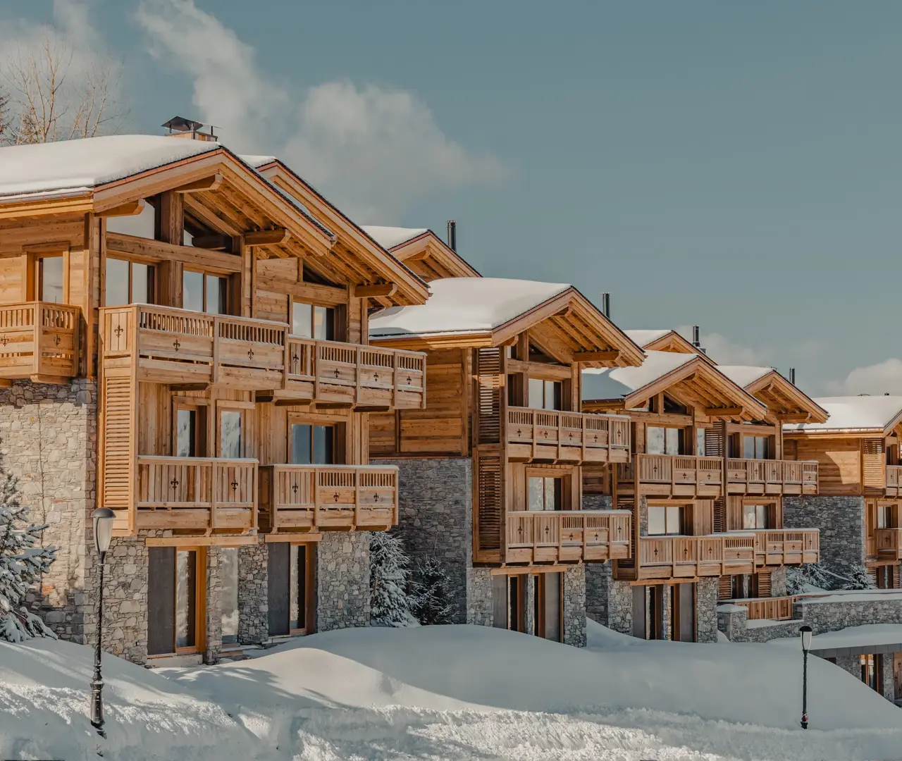 Row of luxury wooden chalets with stone facades covered in snow under a clear blue sky in the mountains.