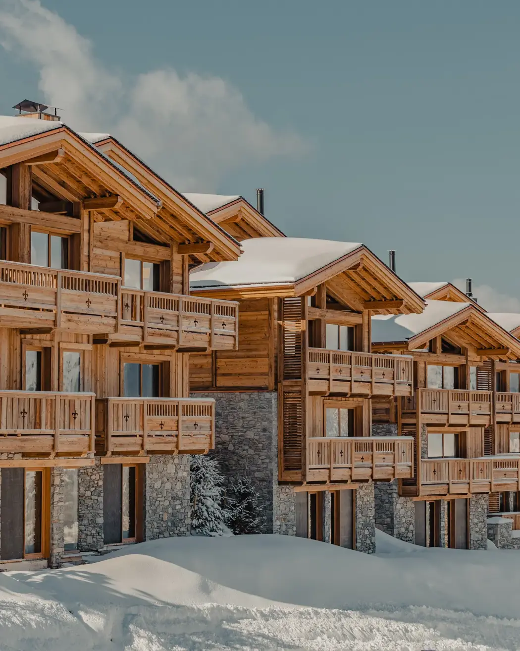 Rangée de chalets de luxe en bois avec façades en pierre, recouverts de neige sous un ciel bleu clair en montagne.