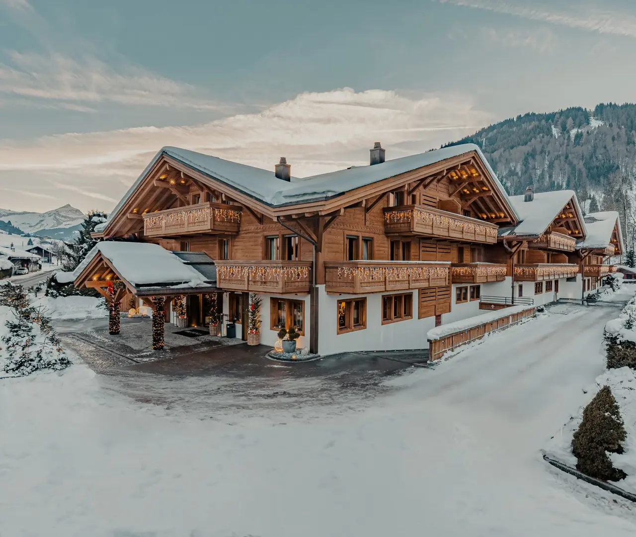 A luxurious alpine chalet surrounded by snow, featuring wooden facades, balconies adorned with warm fairy lights, and a picturesque mountain backdrop under a soft winter sky.