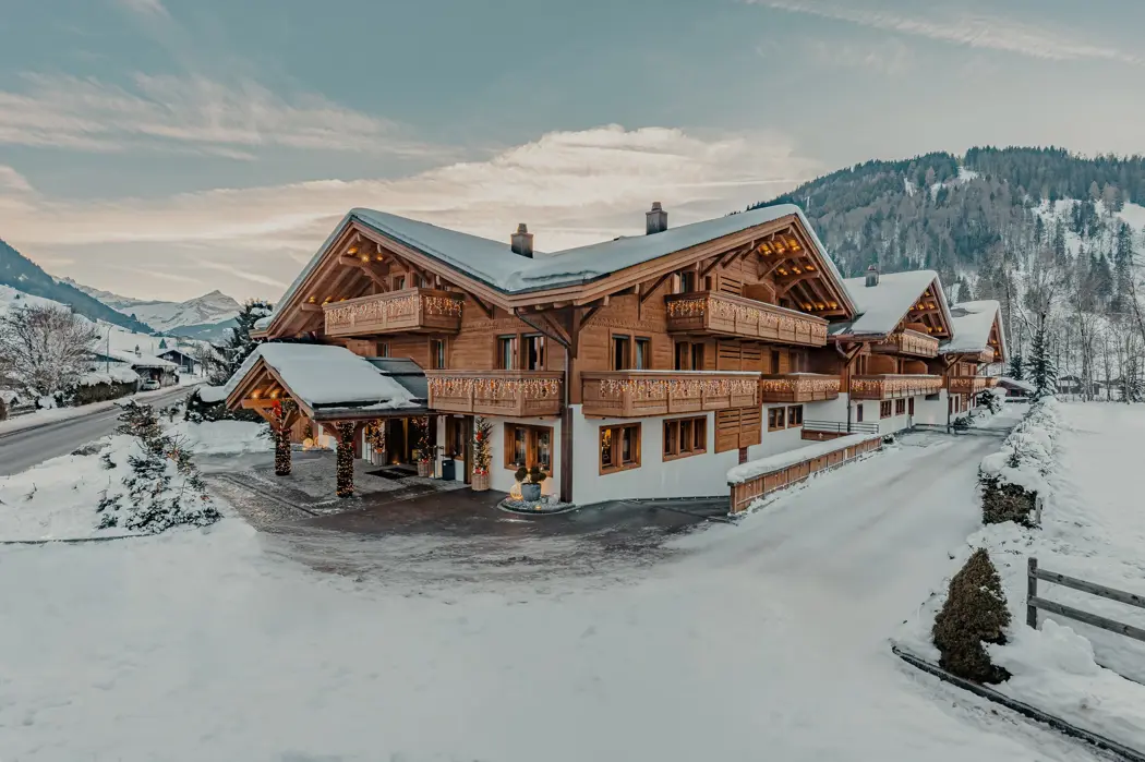 A luxurious alpine chalet surrounded by snow, featuring wooden facades, balconies adorned with warm fairy lights, and a picturesque mountain backdrop under a soft winter sky.