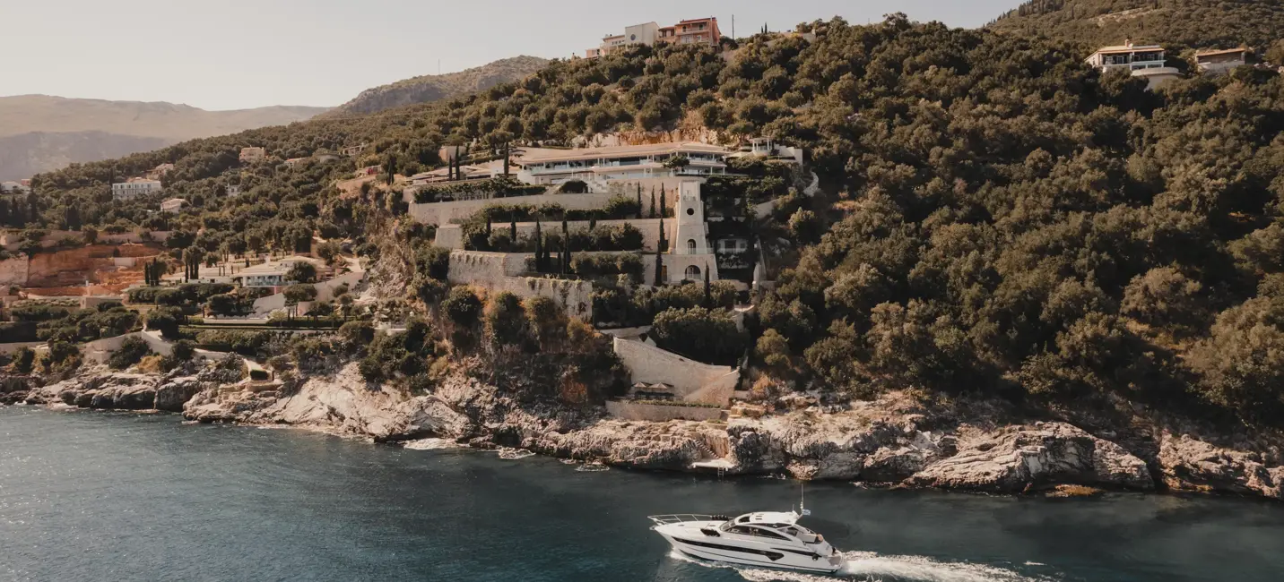 Vue aérienne d’une villa côtière de luxe construite à flanc de colline avec des jardins en terrasses et un yacht naviguant sur la mer turquoise.