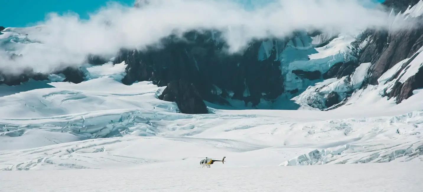Вертолёт стоит на бескрайнем снежном плато у подножия ледниковых вершин, среди облаков и чистого голубого неба.