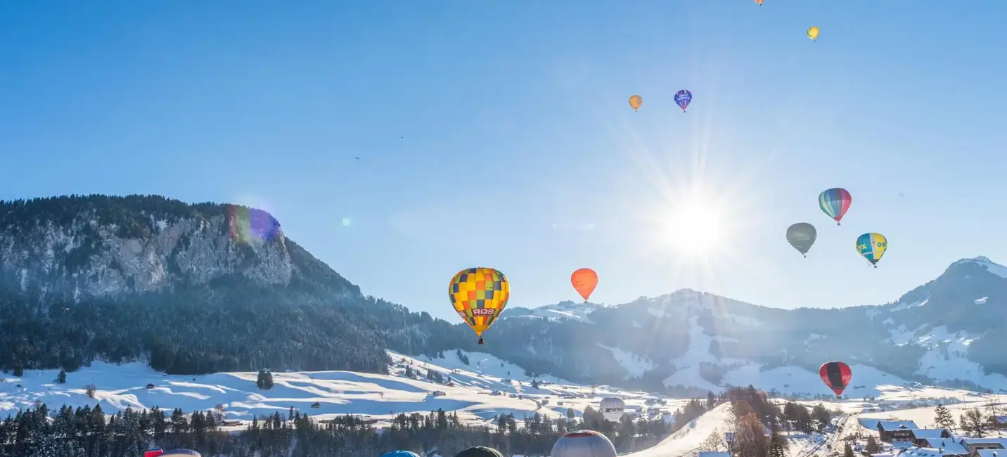 A bright winter scene of colorful hot air balloons floating above a snow-covered alpine valley, with the sun shining over distant mountains and wooden chalets below.