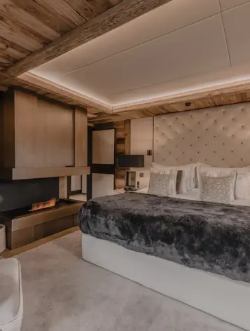 An elegant chalet bedroom with a warm, modern design featuring a large bed with a tufted headboard, soft pillows, and a dark fur throw. The room includes a sleek built-in fireplace, plush armchair, and wooden walls creating a cozy alpine atmosphere.