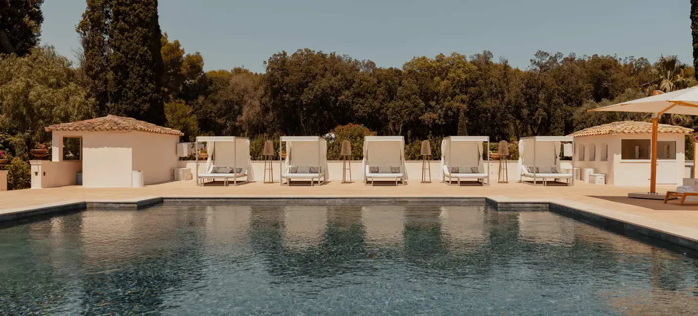 Luxury poolside area with cabanas and sun loungers facing a serene swimming pool, surrounded by greenery and Mediterranean-style pavilions under a clear sky.