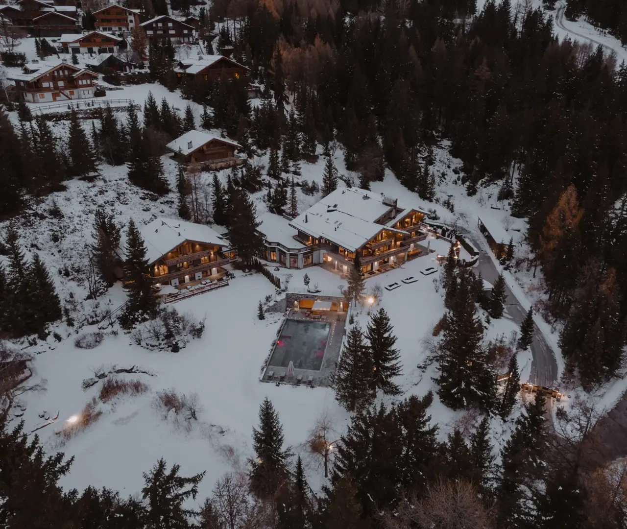 Aerial view of a luxury alpine chalet surrounded by snowy forest at dusk, with warm lights and a heated outdoor pool.”