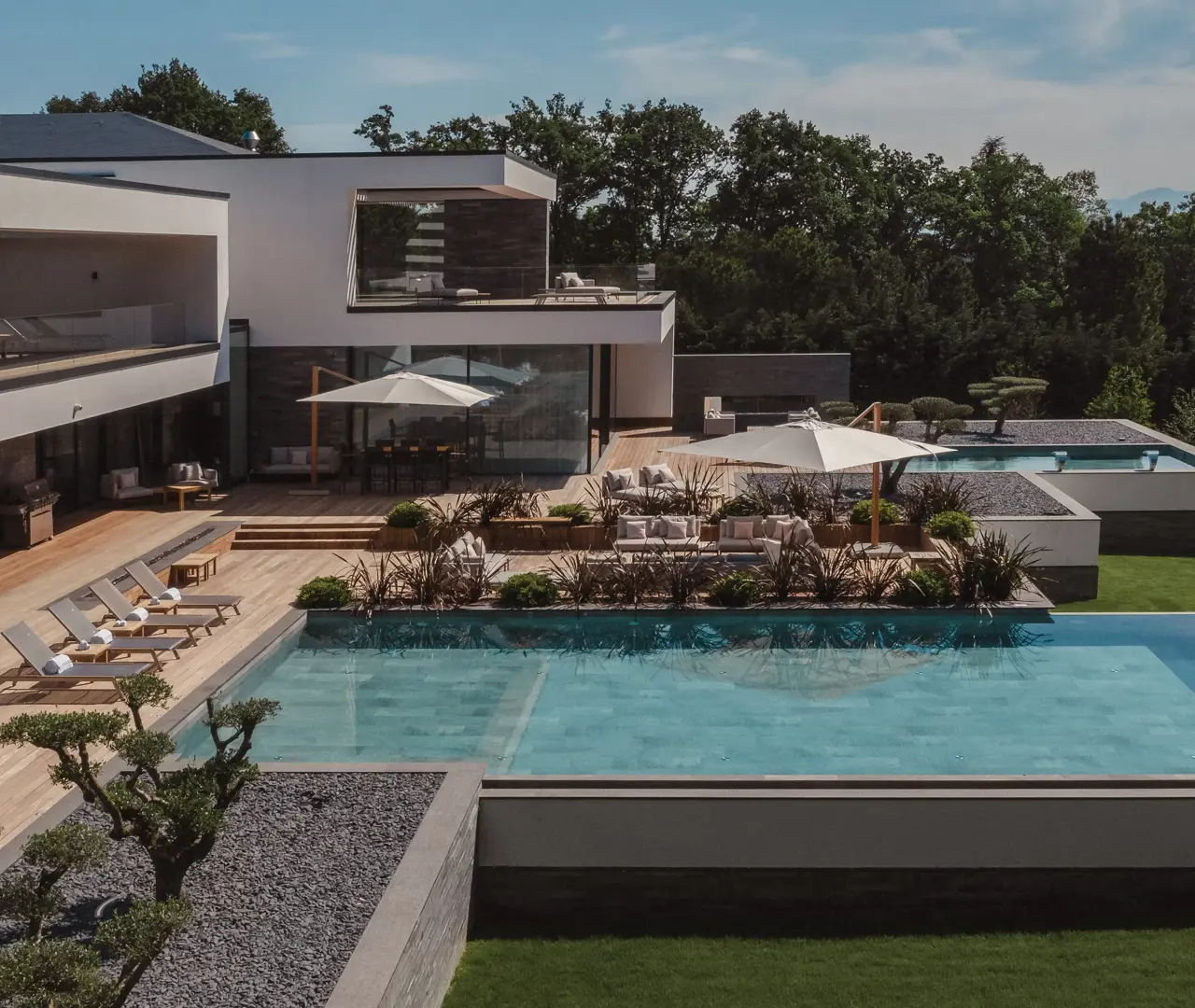 Современная роскошная вилла со стеклянными стенами, просторными террасами и бескрайним бассейном, окружённая зеленью под ясным небом.