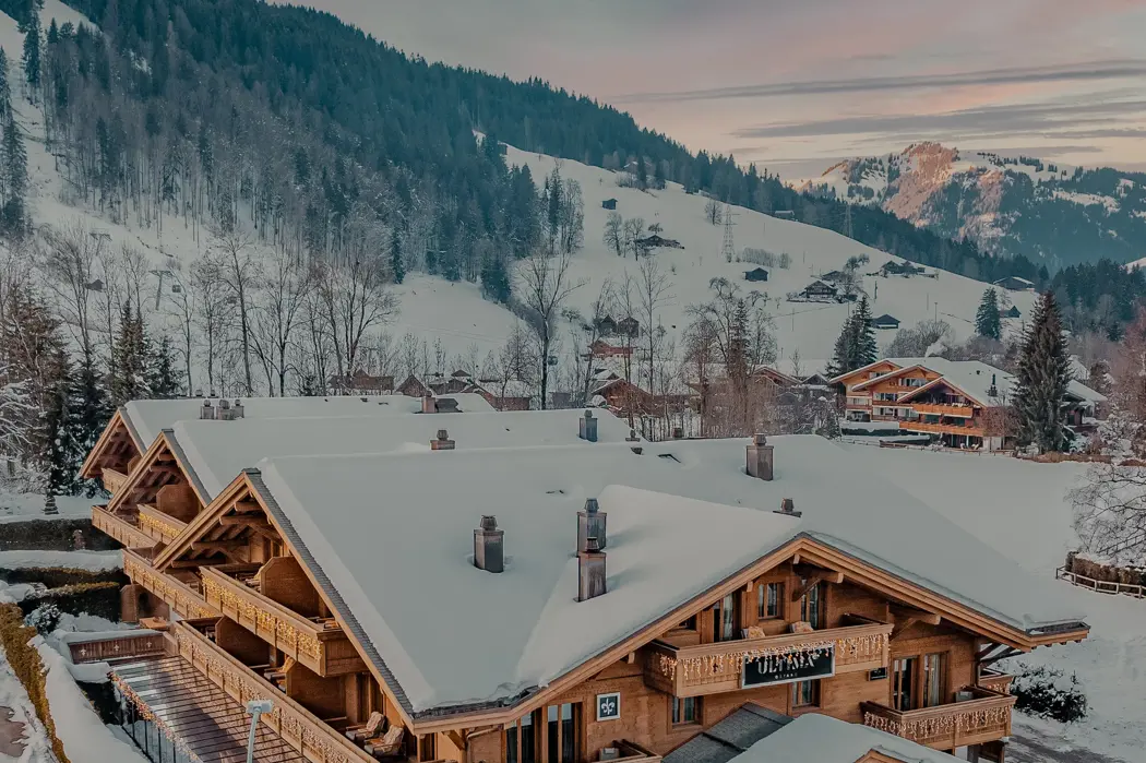 Chalet en bois enneigé entouré de montagnes alpines au lever du soleil, baigné d’une douce lumière dorée illuminant les toits et les pentes boisées.