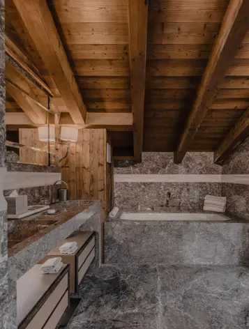 A luxurious bathroom with marble walls and floors, a built-in bathtub beneath a sloped wooden ceiling, and a wide vanity area illuminated by warm lighting through sheer curtains.