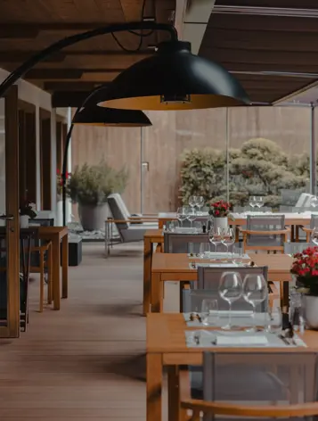Espace de restauration en plein air élégant avec des tables en bois dressées de verres à vin et de nappes blanches, sous des lampes noires modernes et entouré de verdure.