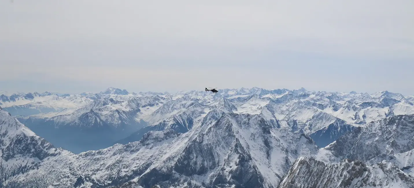 Вертолёт летит над заснеженными альпийскими вершинами в ясный зимний день.
