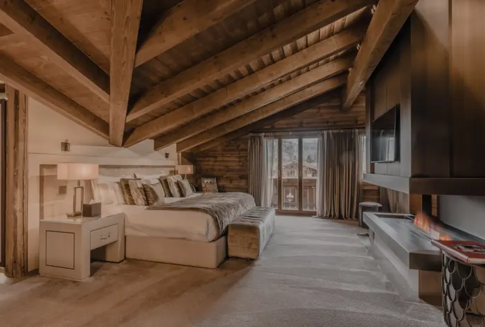 A luxurious chalet bedroom with a sloped wooden ceiling, a large bed dressed in soft linens and fur accents, and a cozy fireplace built into the wall. Natural light pours in through glass doors leading to a balcony with mountain views, creating a warm and inviting alpine atmosphere.