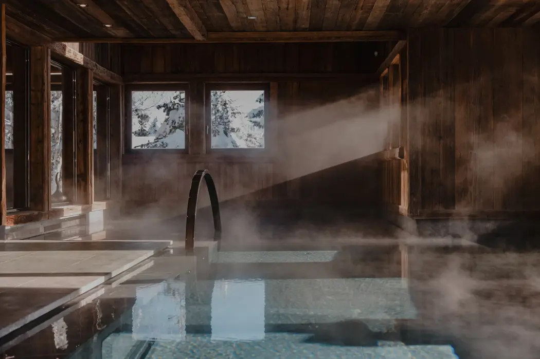 مسبح داخلي فاخر في كوخ جبلي بجدران خشبية وأشعة الشمس تتسلل عبر النوافذ وبخار يتصاعد في أجواء سبا جبلية دافئة.