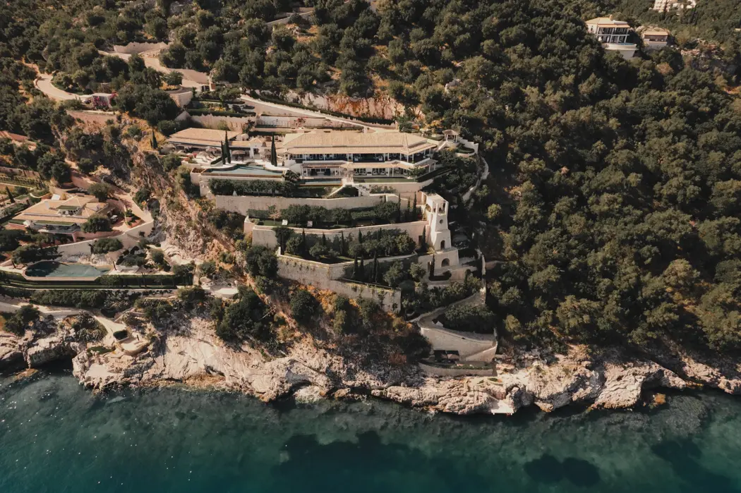 Vue aérienne d’un domaine méditerranéen de luxe perché sur une falaise, entouré de verdure luxuriante, de jardins en terrasses et dominant la mer turquoise.