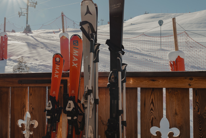 زوج من الزلاجات والعصي يستند إلى سياج خشبي في يوم مشمس على منحدر تزلج مغطى بالثلج، مع شروق الشمس الساطع ومصاعد التزلج في الخلفية.