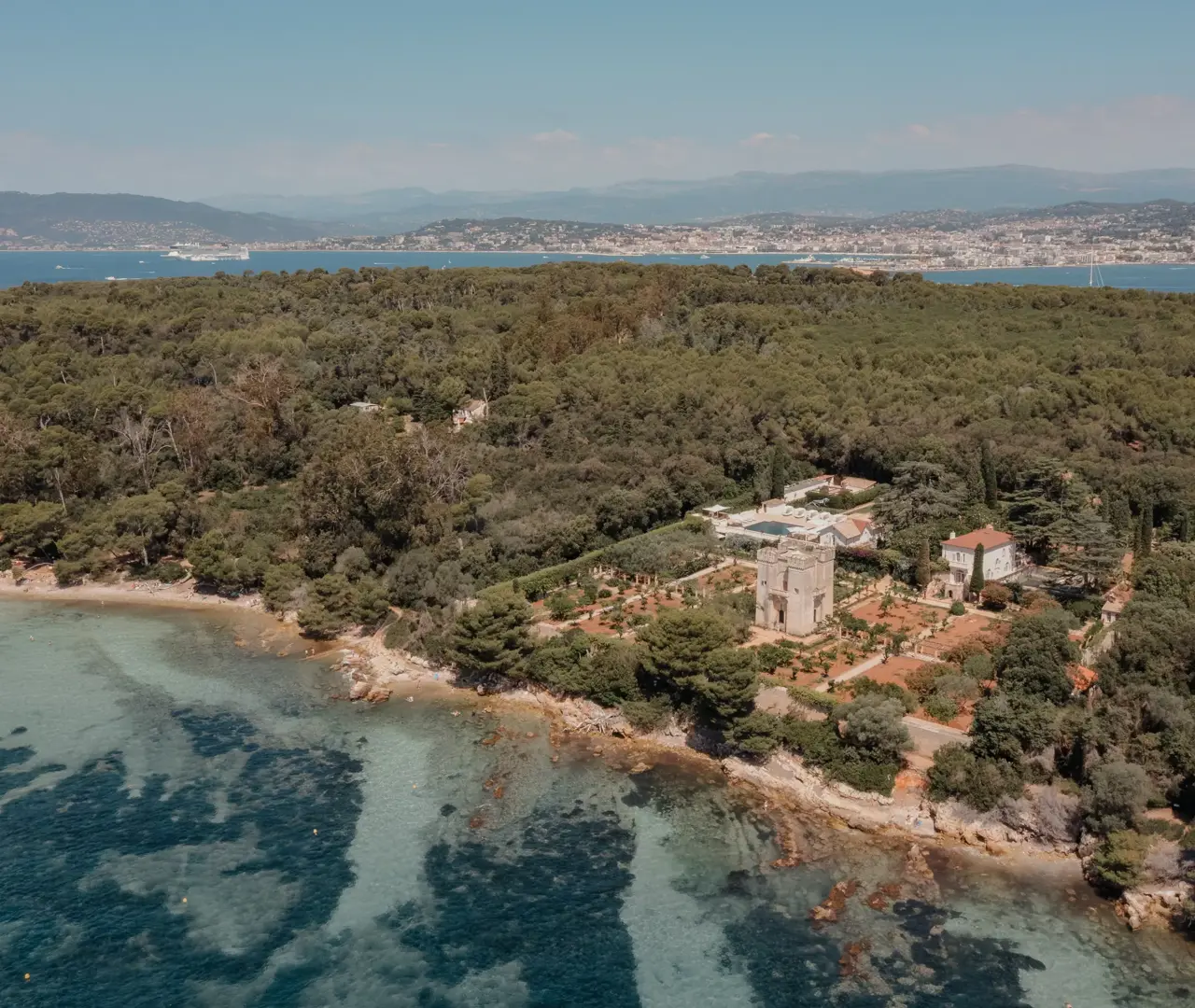 Aerial view of a historic estate surrounded by forest on a Mediterranean island, with turquoise water and a coastal town in the distance.