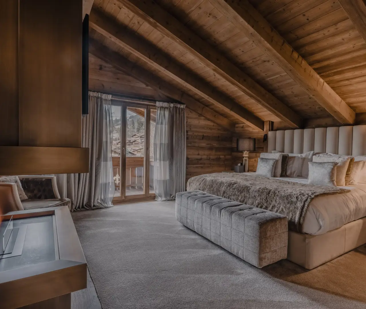Luxurious chalet bedroom with a sloped wooden ceiling, a large bed covered in plush fur throws, a modern fireplace, and glass doors opening to a balcony with mountain views.