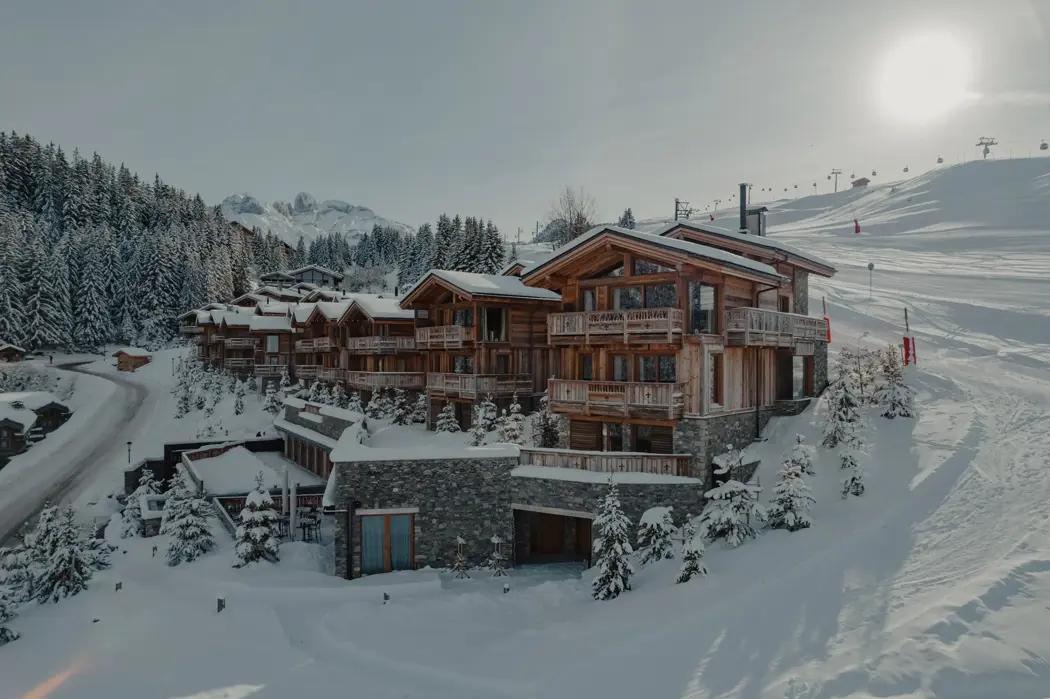 Chalets en bois de luxe nichés dans un paysage alpin enneigé, entourés de pins et de pistes de ski sous un soleil d’hiver éclatant.