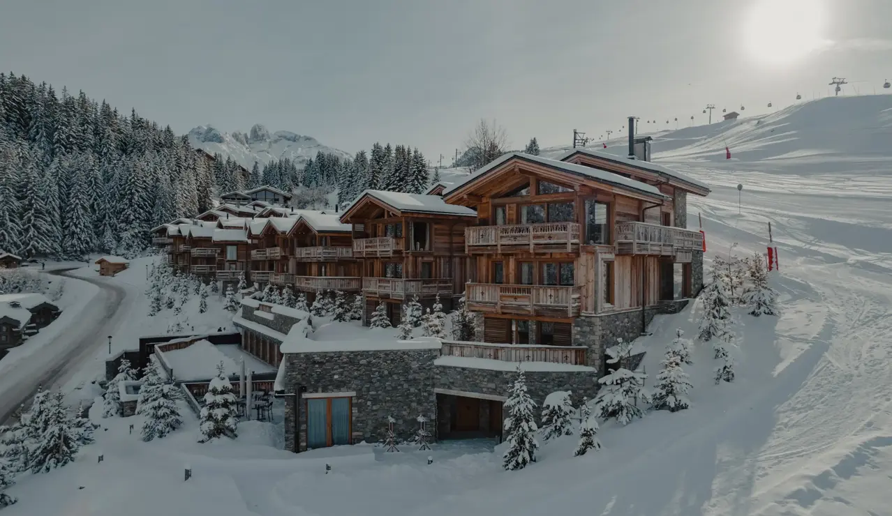Luxury wooden chalets nestled in a snowy alpine landscape, surrounded by pine trees and ski slopes under bright winter sunlight.