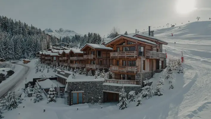 Luxury wooden chalets nestled in a snowy alpine landscape, surrounded by pine trees and ski slopes under bright winter sunlight.