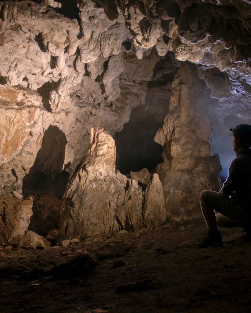 Explorer wearing a helmet and headlamp observing rock formations inside a dimly lit cave.