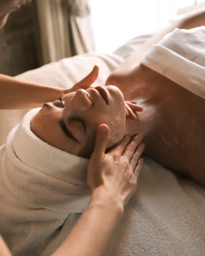 Spa therapist performing a relaxing facial massage on a person wrapped in white towels during a skincare treatment.