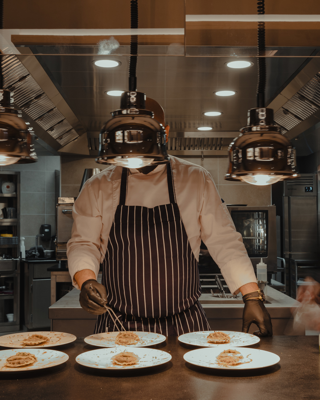 Chef in a professional kitchen plating gourmet dishes under warm pendant lights, with sous-chefs moving in the background.