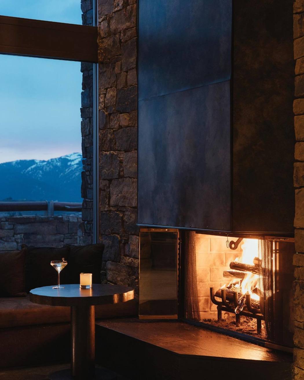 Cozy lounge with stone fireplace, burning fire, and mountain view at dusk.