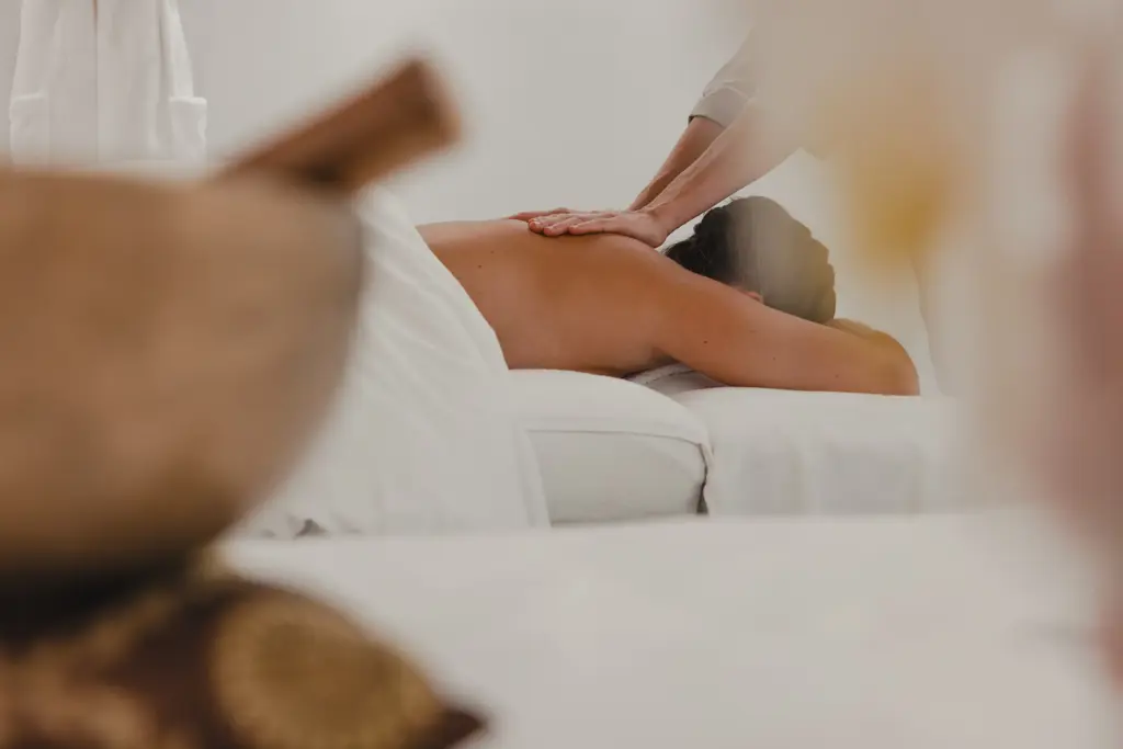 A relaxing spa scene showing a person receiving a back massage on a white treatment bed in a serene, softly lit environment.